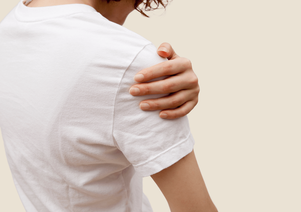 Person in white shirt holds onto their aching shoulder. Beige background.